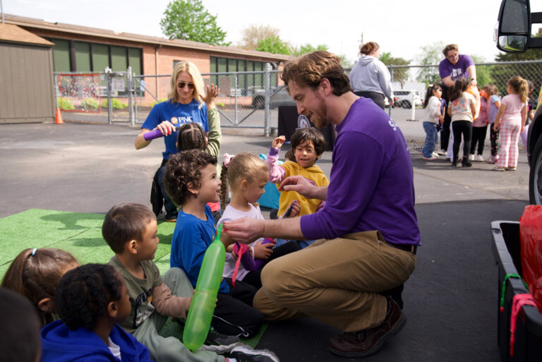 An image of a man in a Science City shirt helping children with a science experiment where they blow up balloons and release them. He is assisted by a woman in a PNC shirt.